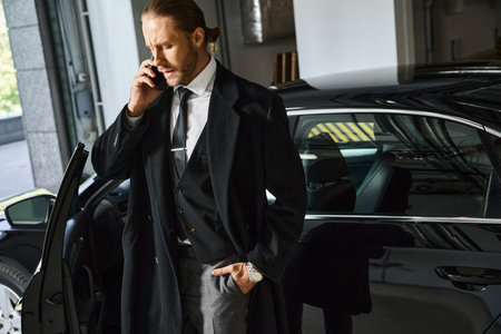 good looking elegant man in black coat talking by phone on parking lot with hand in pocket, businessの写真素材