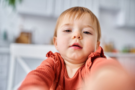 close up of pretty baby girl in orange sweater trying to reach out actively camera with her hands