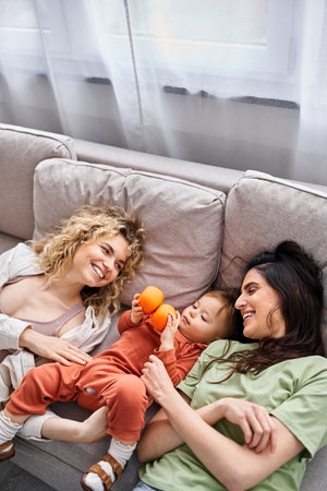 joyous lgbt couple lying on sofa with their pretty baby girl holding tangerines, family concept