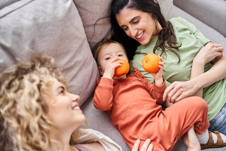 cheerful lgbt couple lying on sofa with their pretty baby girl holding tangerines, family conceptの写真素材