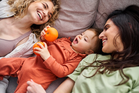 beautiful happy lgbt couple lying on sofa with their baby girl holding tangerines, family concept