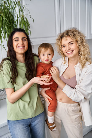 caring cheerful lesbian couple in homewear posing happily with their baby girl, modern parenting