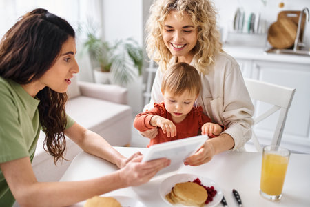 beautiful lgbt couple having breakfast with their baby girl and looking at tablet, family conceptの写真素材
