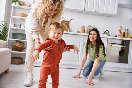 cheerful lesbian couple in homewear helping their adorable baby girl to stand, family conceptの写真素材