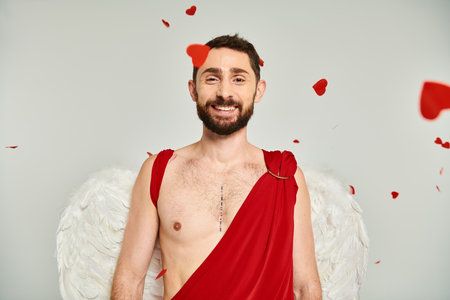 bearded man in cupid costume and wings smiling at camera under red heart-shaped confetti on greyの写真素材