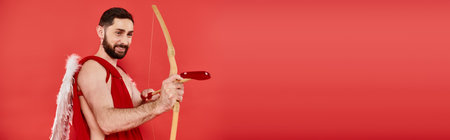 joyful bearded man dressed as cupid shooting from bow with heart-shaped arrow on red, bannerの写真素材