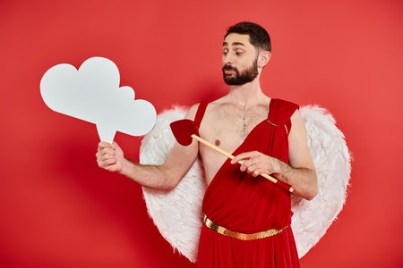 pensive cupid man with heart-shaped arrow looking at white empty thought-bubble on red backdropの写真素材