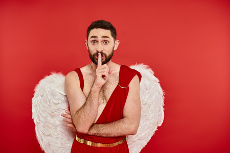 excited bearded man in cupid costume showing shh sign and looking at camera on red, st valentinesの写真素材