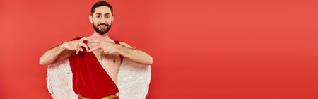 cheerful bearded man dresses as cupid showing heart sign and looking at camera on red, banner