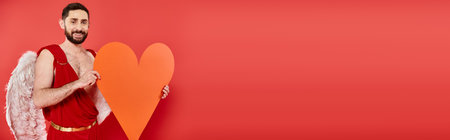 whimsical bearded man in cupid costume with huge orange paper heart on red, horizontal bannerの写真素材