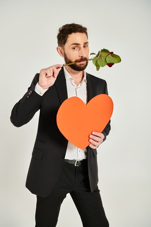 bearded stylish man with orange paper heart and red rose in teeth on grey, st valentines conceptの写真素材