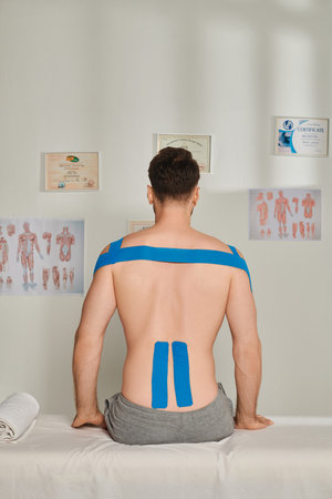 back view of man sitting during appointment with kinesiological tapes on his back, healthcare