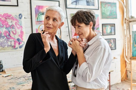 fashionable lesbian couple holding hands and looking around in workshop with creative paintingsの写真素材