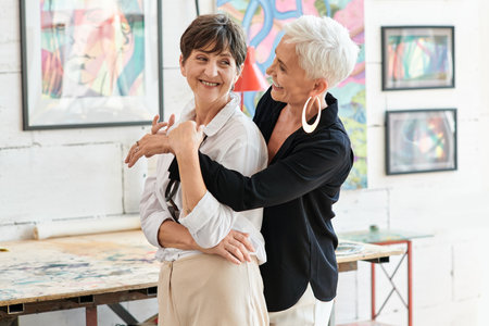 cheerful and trendy mature woman embracing lesbian partner in modern art workshop, happinessの写真素材