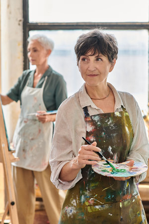 smiling mature woman holding color palette and looking away during master class in art workshopの写真素材