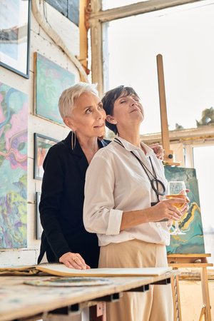 mature woman with closed eyes and wine glass near stylish lesbian partner in contemporary art studioの写真素材