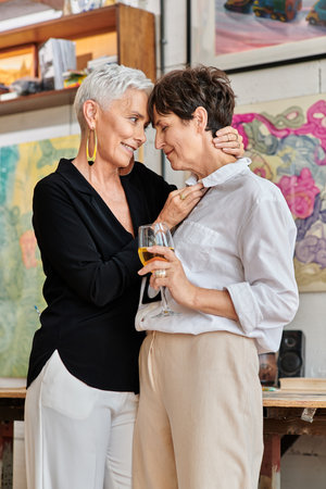 stylish middle aged woman embracing lesbian partner with wine glass in contemporary art studioの写真素材
