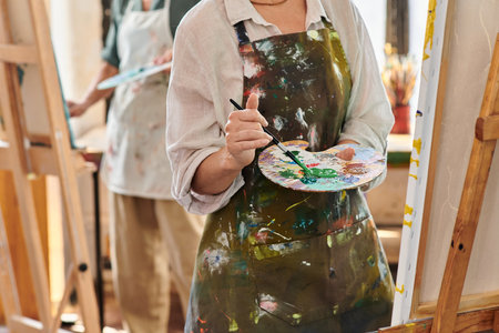 cropped view of mature woman in apron mixing paints on color palette, master class in art workshopの写真素材