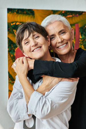 portrait of happy and trendy mature lesbian artists smiling at camera in modern craft workshopの写真素材