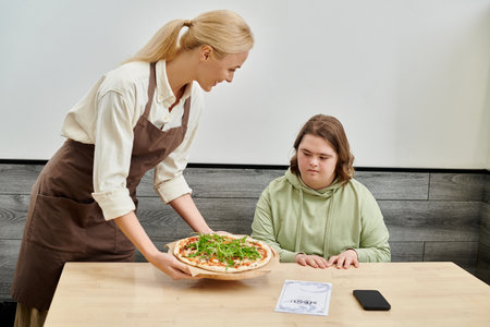 smiling waitress proposing delicious pizza to female client with down syndrome sitting in cozy cafeの写真素材