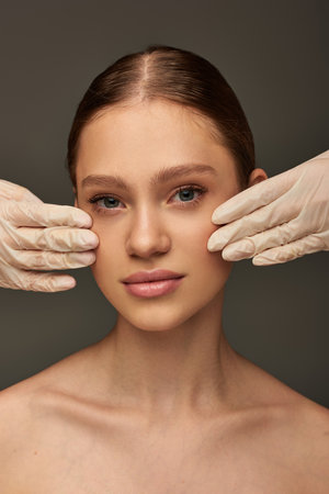 estheticians in medical gloves touching face of pretty woman on grey background, dermatology conceptの写真素材