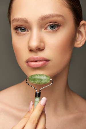 young woman doing face massage with green jade roller on grey background, skin rejuvenationの写真素材