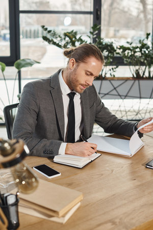 concentrated businessman in formal wear working with notebooks near smartphone with blank screenの写真素材