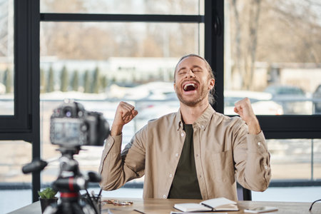 overjoyed businessman showing win gesture in front of digital camera during video blog in officeの写真素材