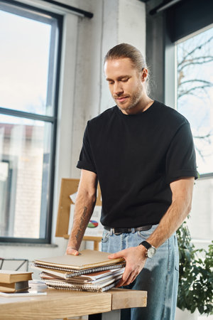 stylish businessman in casual attire holding pile of notebooks while standing at work desk in office