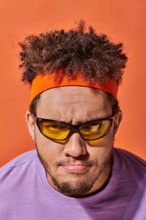 young african american man in eyeglasses and headband looking at camera on orange background,...