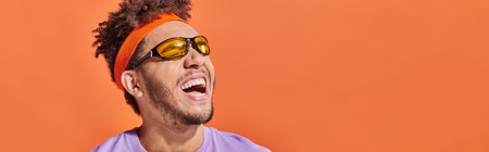 african american man in sunglasses and headband laughing and looking away on orange backdrop, bannerの写真素材