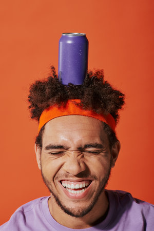 purple soda can on head of happy curly african american guy with headband on orange backgroundの写真素材