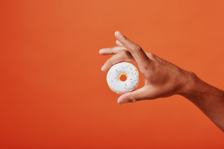 cropped shot of person holding glazed vanilla donut with sprinkles on orange background, white icingの写真素材