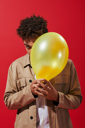 young african american man in beige jacket hiding behind balloon on red background, obscuring faceの写真素材