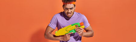african american man in headband playing water fight with toy gun on orange background, banner