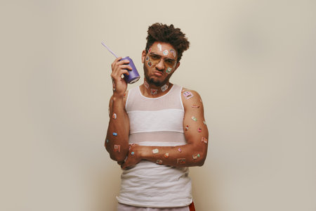 sad african american man with stickers on face and body holding soda can on grey background