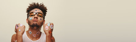 african american man in sunglasses with stickers on face looking at camera on grey backdrop, banner