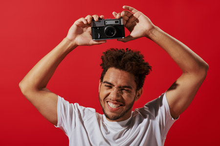joyful african american photographer holding retro camera and smiling on red backgroundの写真素材
