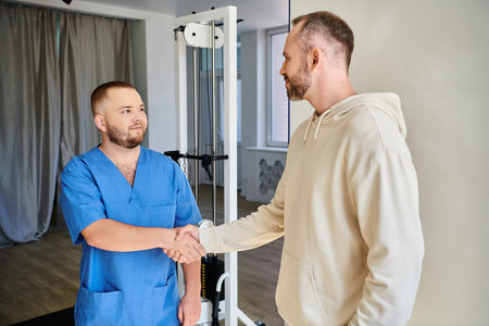bearded man shaking hands with smiling doctor in blue uniform in rehabilitation kinesio centerの写真素材