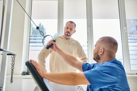 man looking at professional physiotherapist training on exercise machine in kinesiology center