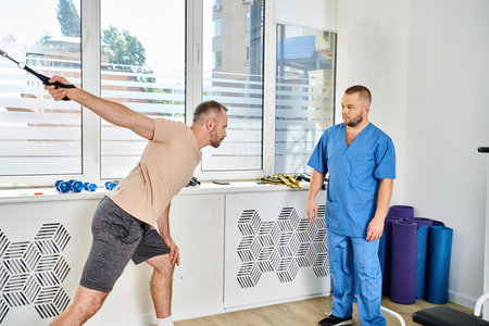 man in sportswear training on exercise machine near experienced rehabilitologist in kinesio center