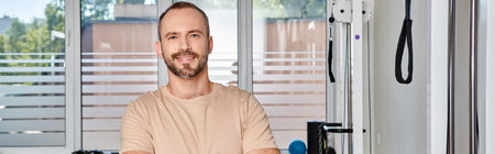 cheerful bearded man with folded arms looking at camera in gym of modern kinesiology center, banner