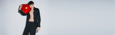 young man in black shiny open shirt holding red balloon on grey background, Valentines day bannerの写真素材