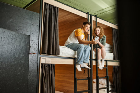 cheerful young woman showing smartphone to boyfriend on double-decker beds in cozy hostel room