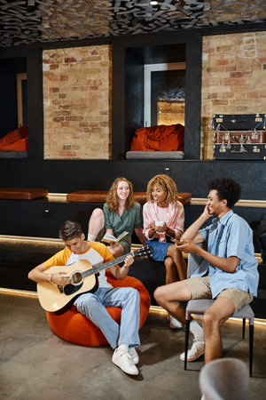 young man playing guitar to cheerful multiethnic friends in lounge of students hostel, travelers
