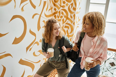 cheerful interracial female friends with paper cups and backpacks on staircase of modern hostelの写真素材