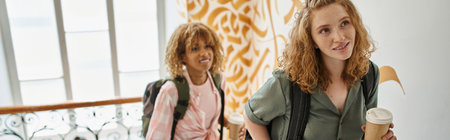 happy interracial female friends with paper cups walking on hostel staircase, horizontal banner