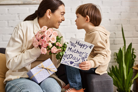 pretty jolly mother posing on sofa with her toddler son with flower bouquet and present, Mothers dayの写真素材