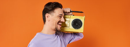 banner of happy man in purple sweatshirt holding boombox and listening music on orange background