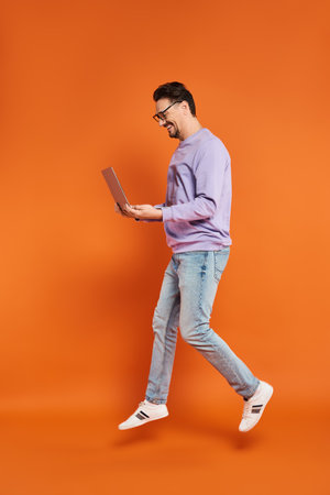 cheerful man in eyeglasses and purple sweater using laptop on orange background, remote workの写真素材